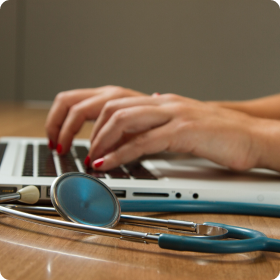 A closeup of a person typing on a laptop, with a stethoscope lying next to the laptop on the desk